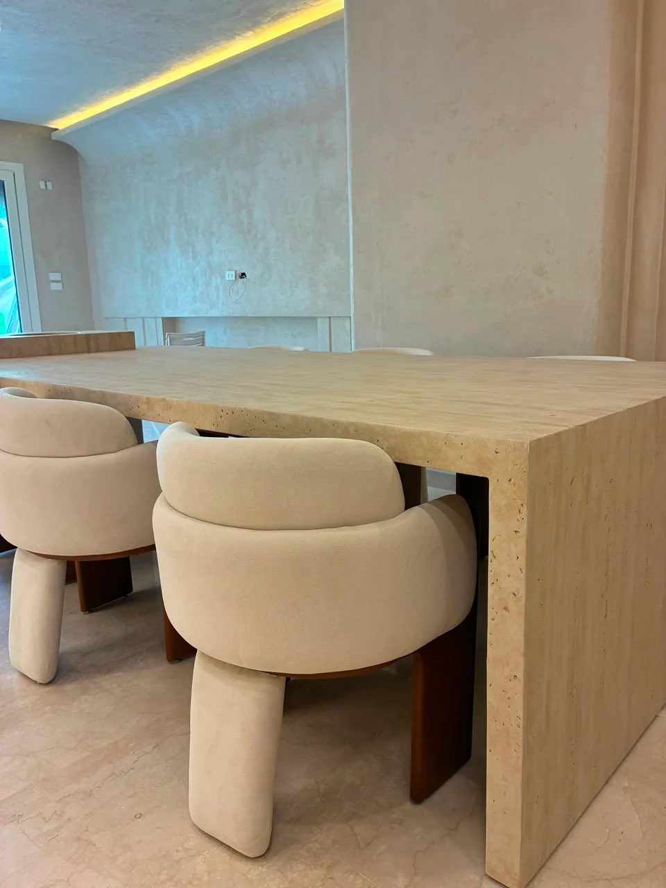 A travertine dining table with cream upholstered dining chairs on wooden legs, photographed in a finished room